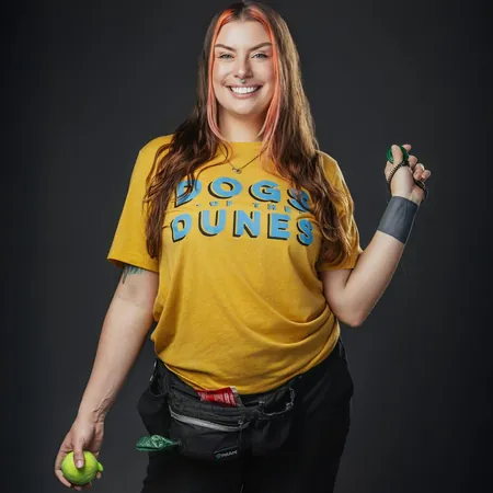 Theresa Armstrong is wearing a yellow Dogs of the Dunes shirt, holding a dog training tool in one hand and tennis ball in the other. A fanny pack containing dog training equipment is around her waist. She is smiling widely, ready to help customers with all of their dog training needs.